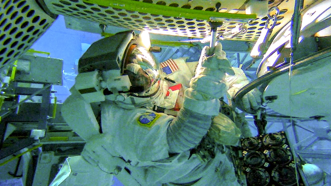 A view of NASA astronaut Victor Glover in the Neutral Buoyancy Lab at Johnson Space Center, wearing a space suit and helmet. He tests a tool for swabbing the exterior of space station. Various mock spacecraft parts used for training are submerged underwater in the background. The astronaut's face is vaguely visible in the helmet visor, showing the bright lights of the spacecraft.