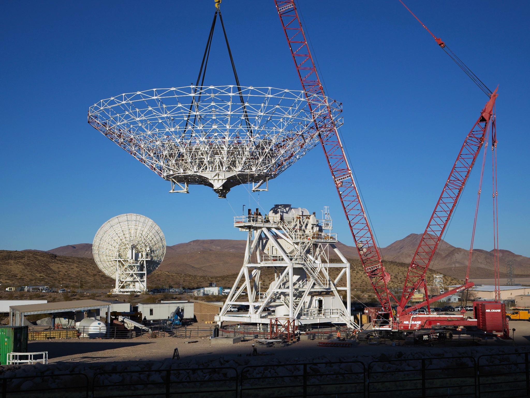 A large red crane lowers a cone-shaped frame. There is a large white metal structure behind it that has stairs running around it, all the way to the top. In the background at left, a completed antenna faces away from us. Farther away in the background are brown mountains.
