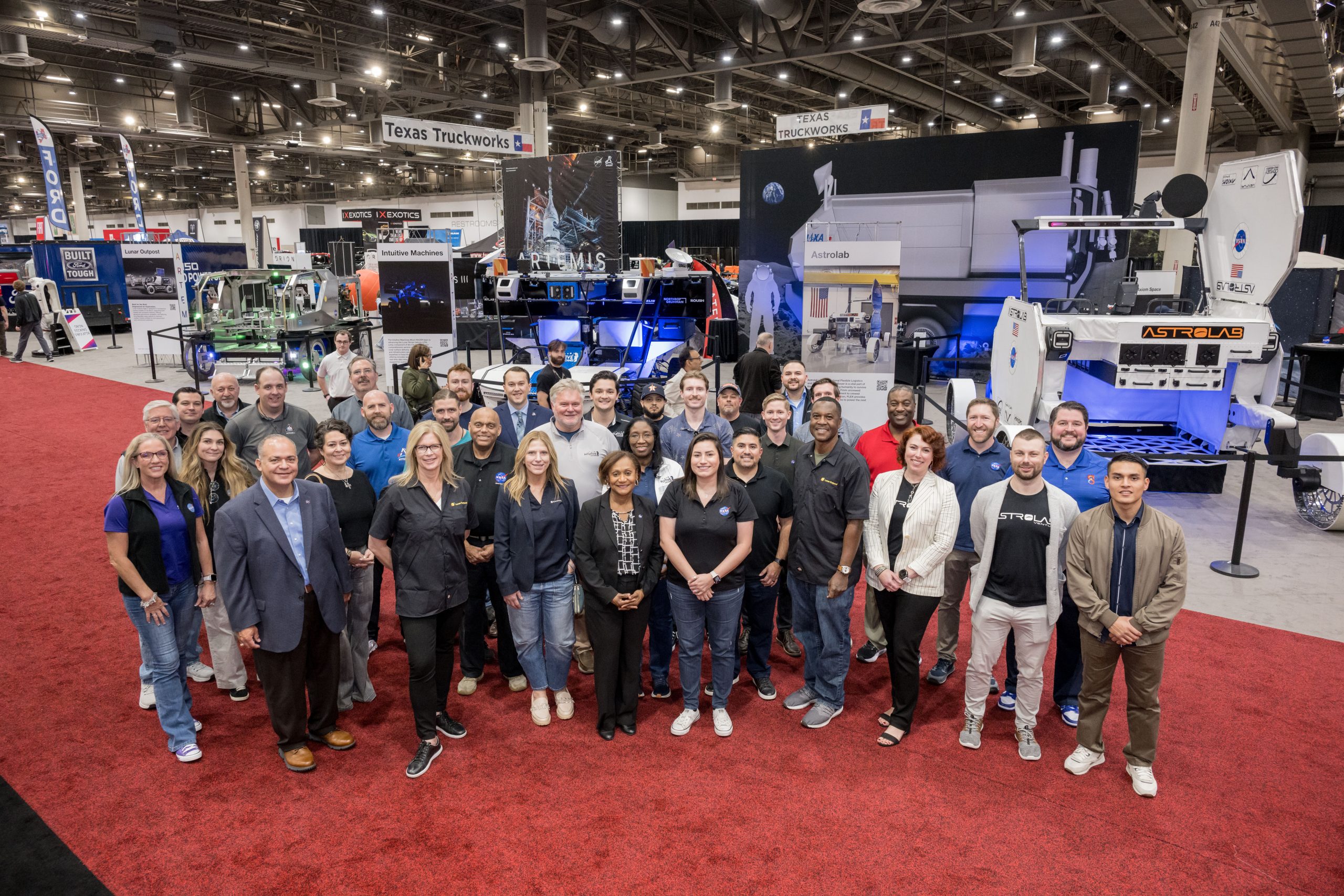 Group photo of individuals standing on a red carpet at a convention center, surrounded by exhibits showcasing space exploration technology.