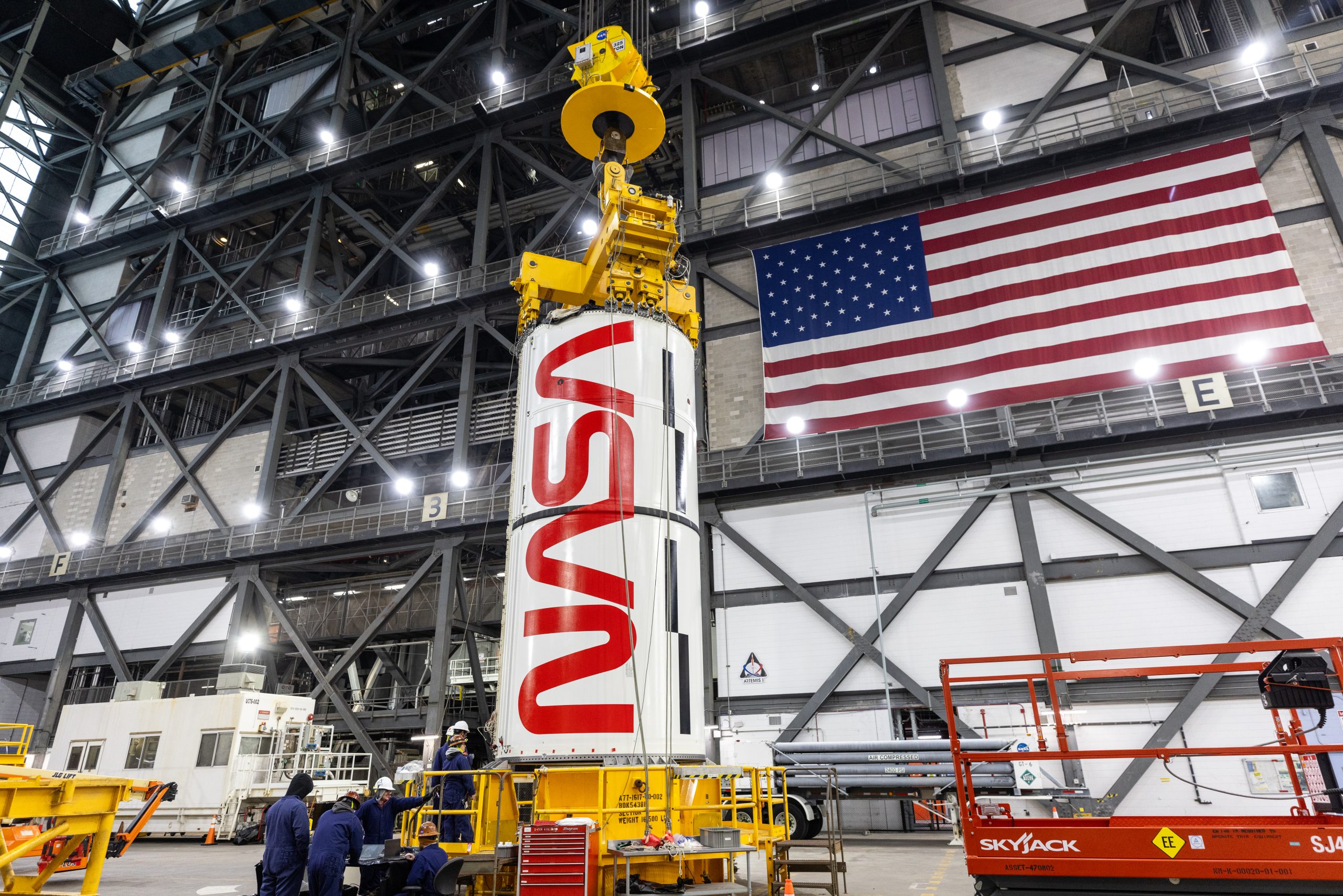 A booster segment - a massive white cylindrical object with the red NASA "worm" insignia on it - is attached to a yellow lifting beam inside a large building. There is a large United States flag on the wall to the right of the booster segment. A few people in blue jumpsuits and hard hats stand at the base of the yellow platform at center.
