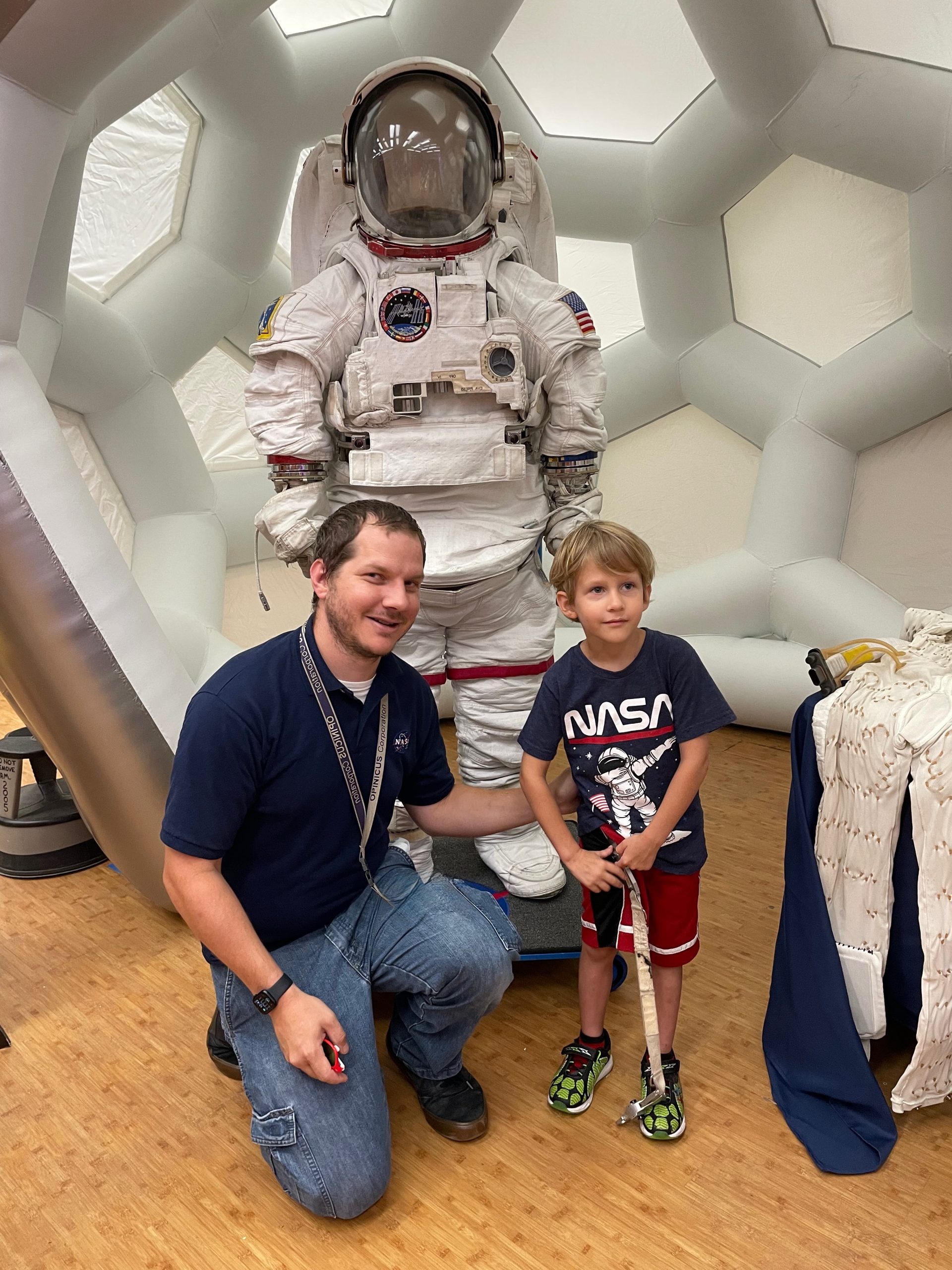 A man, left, kneels down next to a boy with a mockup of an astronaut spacesuit behind them.