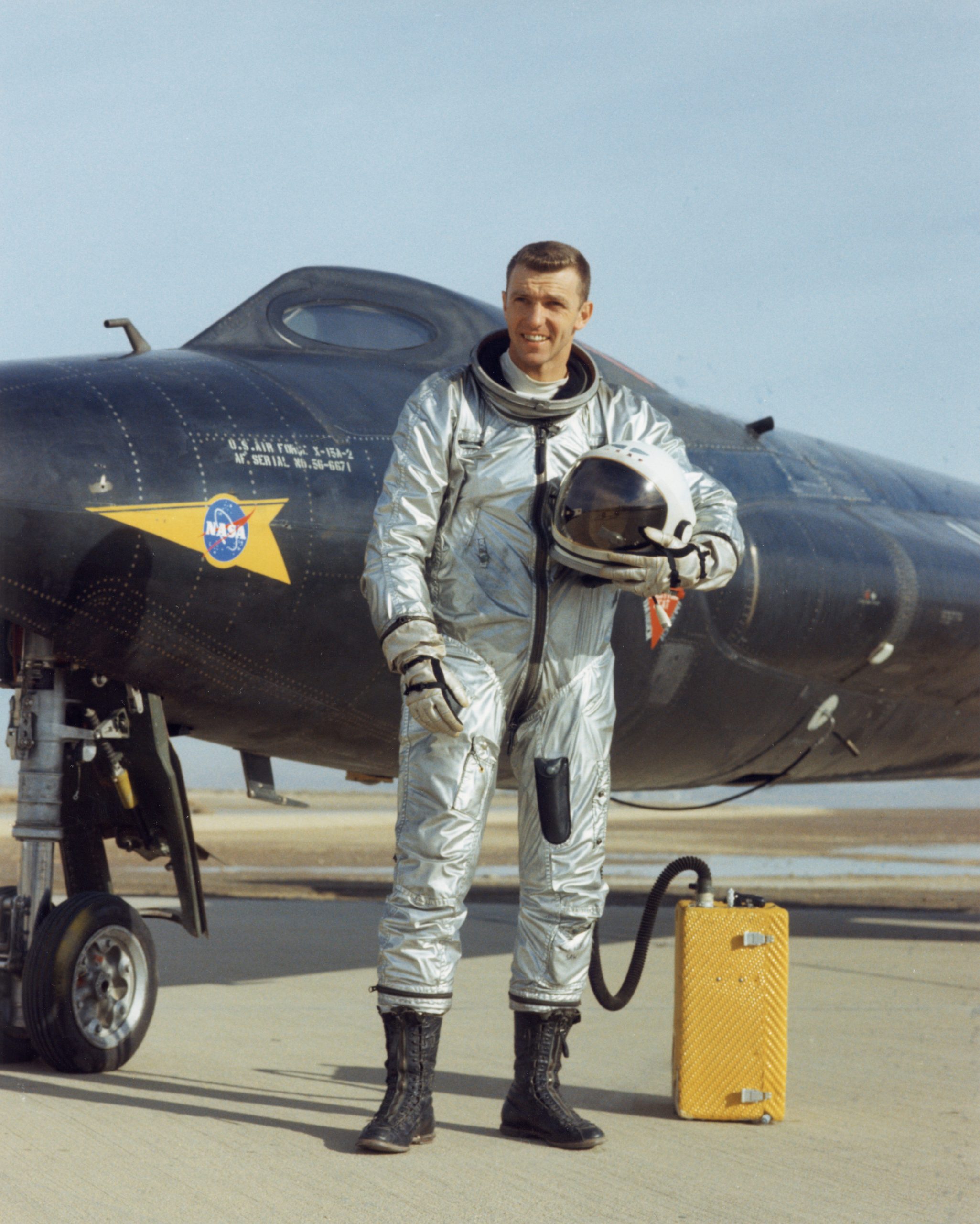A man in a silver suit holds a helmet in his left arm. He looks off to the side. He is standing in front of a black plane.