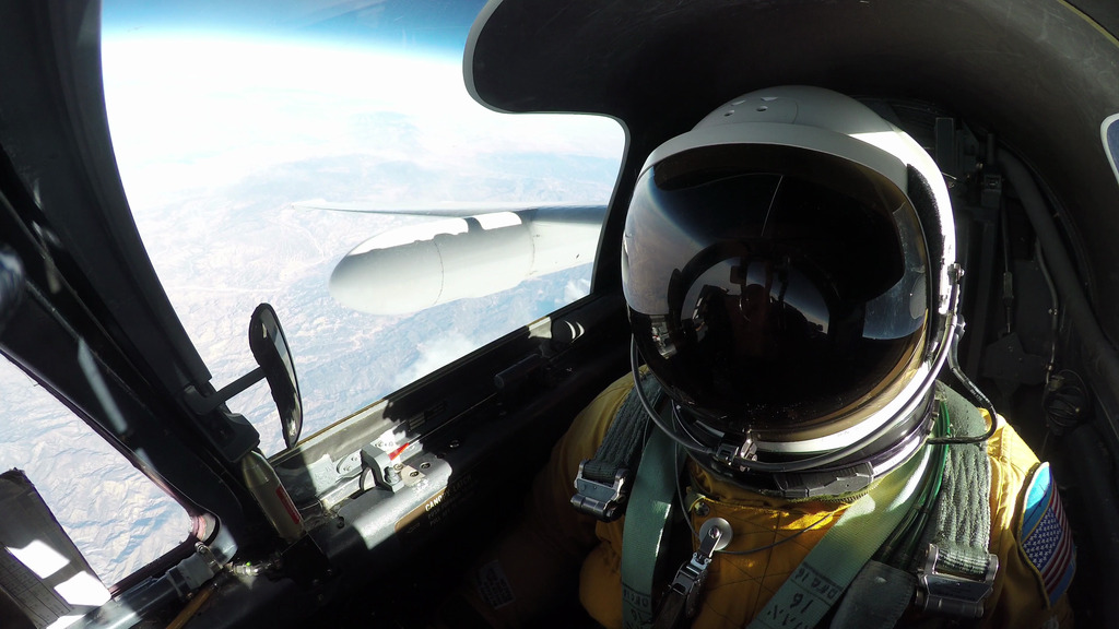 A high-angle, eye-level shot from inside the cockpit of an ER-2 aircraft shows a pilot in a full pressure suit and helmet, with the right wing of the plane and the Earth's horizon visible outside the window. The pilot's helmet is white and reflective, with a dark visor that reflects parts of the cockpit. The pilot is wearing a yellow flight suit and green harnesses.