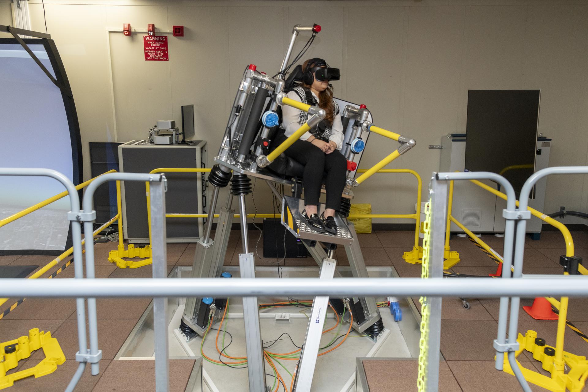 A woman sits in a seat with straps atop a metal platform that is tilted to the right side with three base pieces. Her feet rest on a metal platform. Yellow metal bars surround the platform. The woman is wearing black pants and a black and white vest, as well as black virtual reality goggles and a headset.