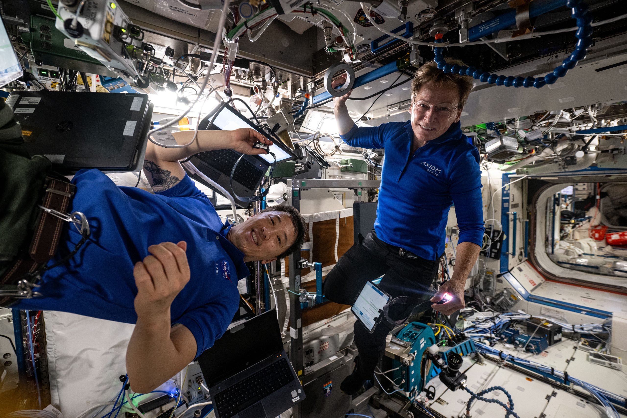 An Asian man and a white woman float inside the International Space Station's Destiny laboratory module. They are both wearing blue shirts and both are looking at the camera. The laboratory module has a lot of exposed cables and parts visible. There are even laptop computers floating nearby.