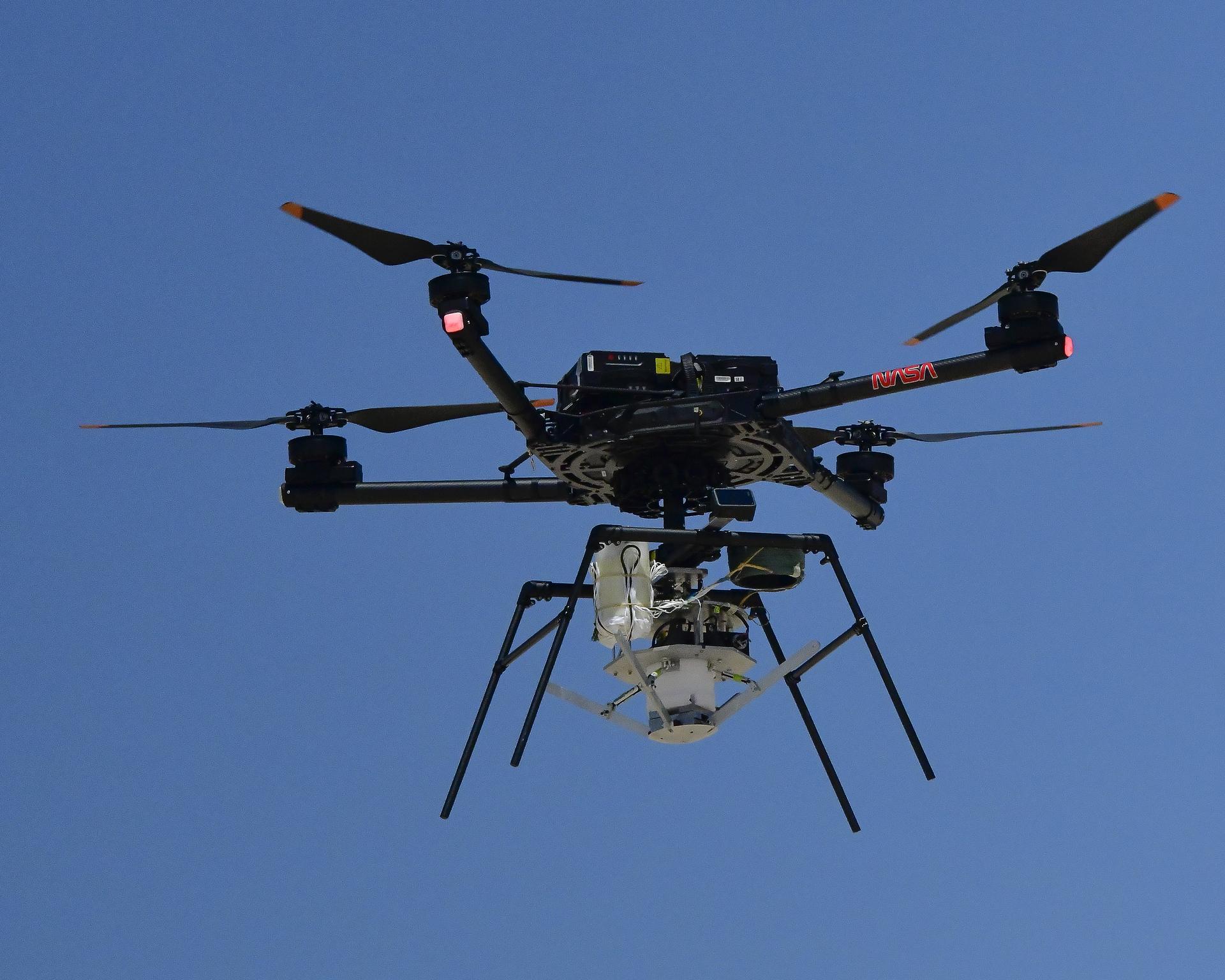 A drone with four rotors hoovers against a canvas of deep blue sky prior to releasing the experiment it carries high above the desert floor.