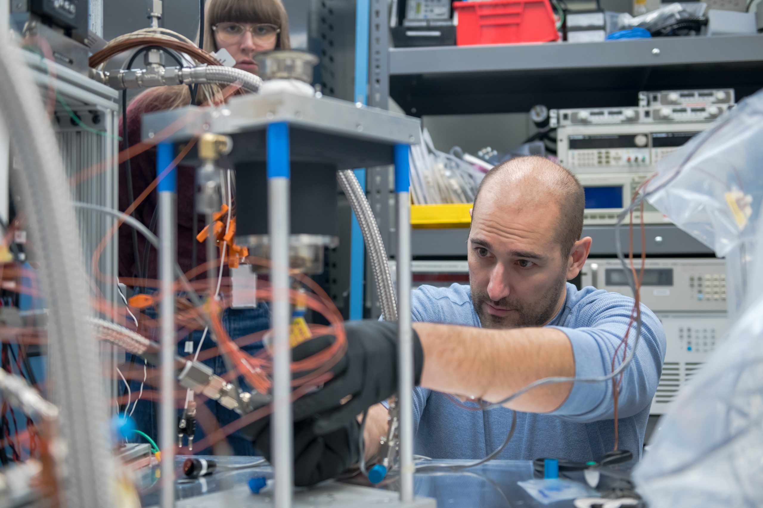 A man wearing a blue shirt leans over a tabletop to make adjustments to a Stirling engine testbed. The mechanism showcases three pillars, about 12 inches in height with a metal cover. Orange winding cords are visible.