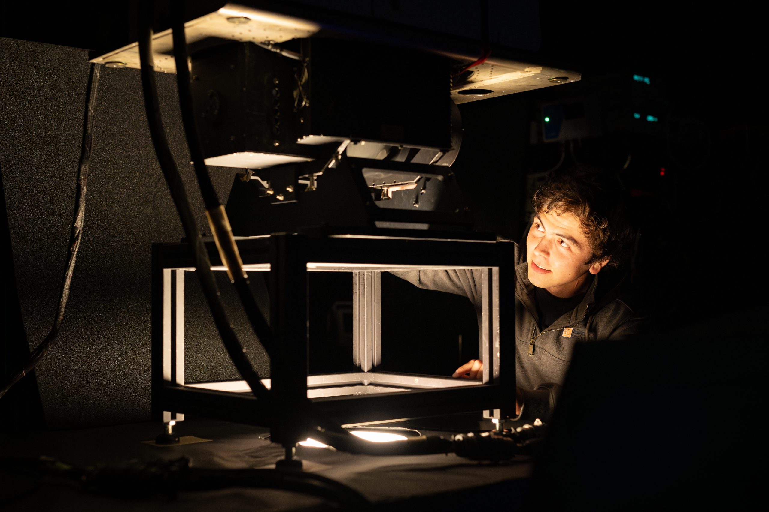 A photo of a room that is mostly dark, with a single light source coming out of a hole in a table. A young engineer with short dark hair looks at the instrument mounted on top of the table, with just his face illuminated by the light.
