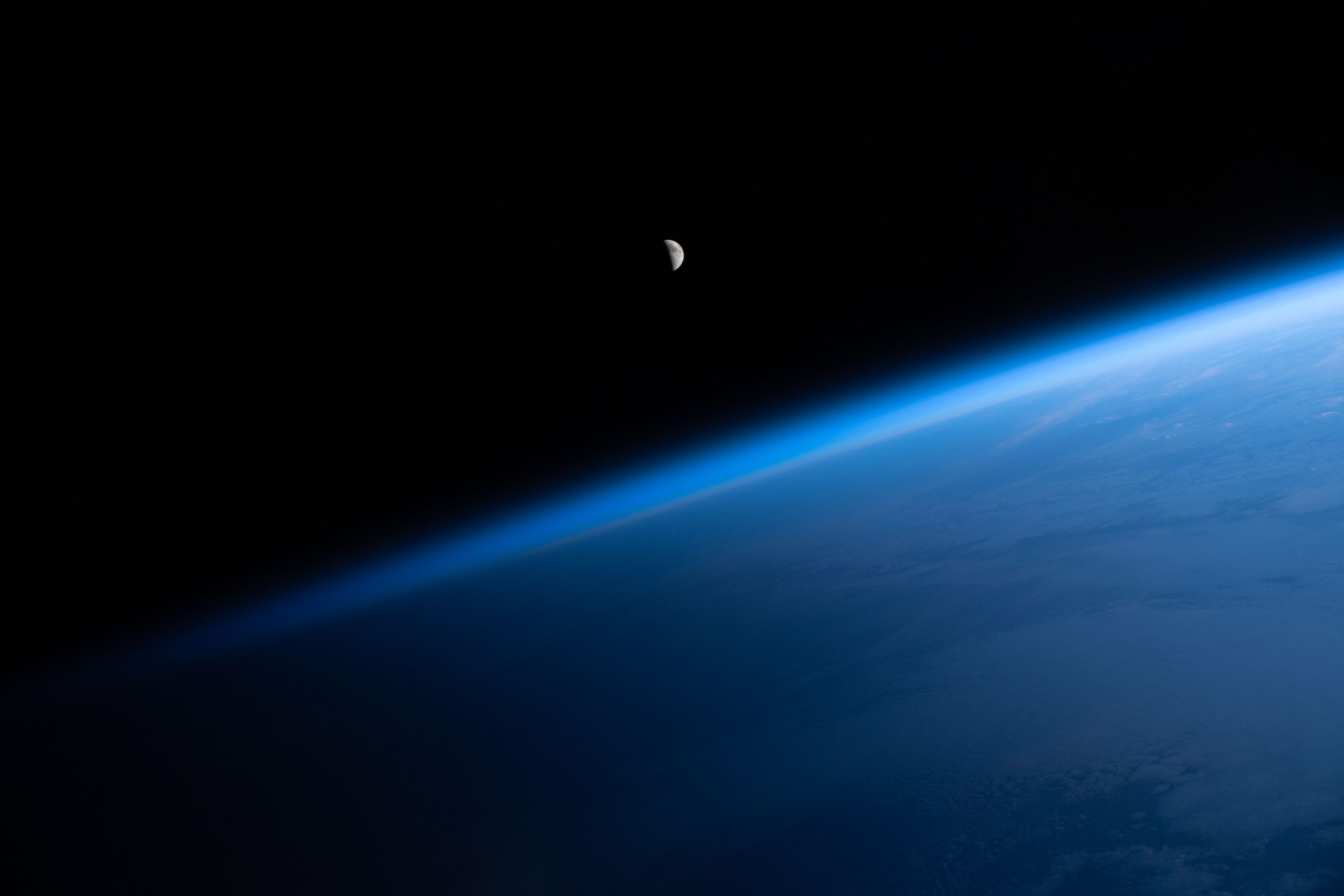 The Moon is a small gray circle with less than half of it visible. Below it, Earth's atmosphere is a brilliant blue haze making a diagonal line across the image.