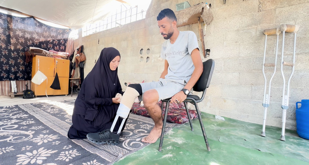 Ibrahim Abdel Nabi, a Palestinian displaced in Gaza, sitting on a chair while his wife helps him wear the handmade prosthetic limb. Ibrahim Abdel Nabi, a Palestinian displaced in Gaza, sitting on a chair while his wife helps him wear the handmade prosthetic limb.