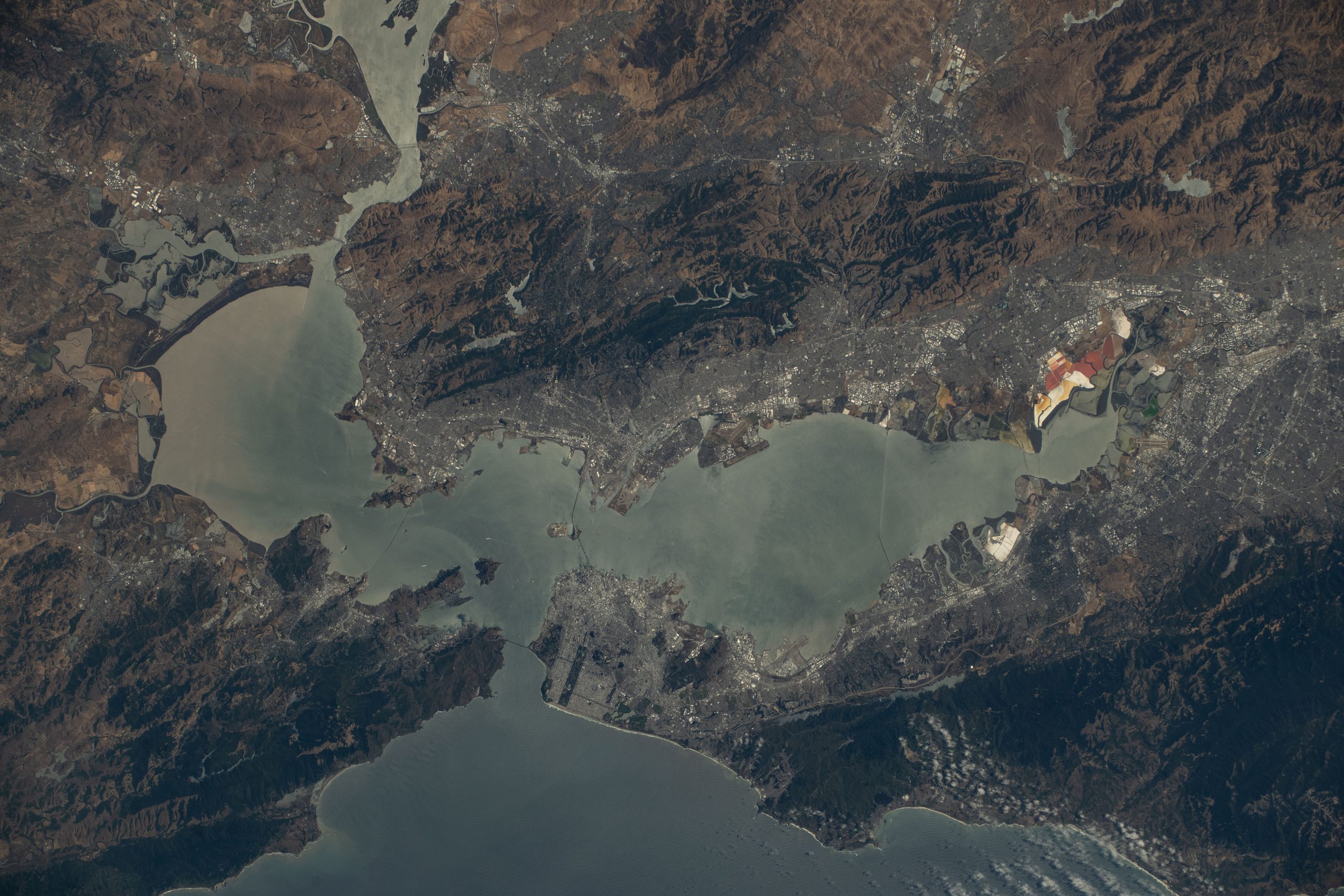 California's San Francisco Bay Area as seen from the International Space Station orbiting 260 miles above. The water is slate gray, while the surrounding land is in shades of brown, deep green, and gray.