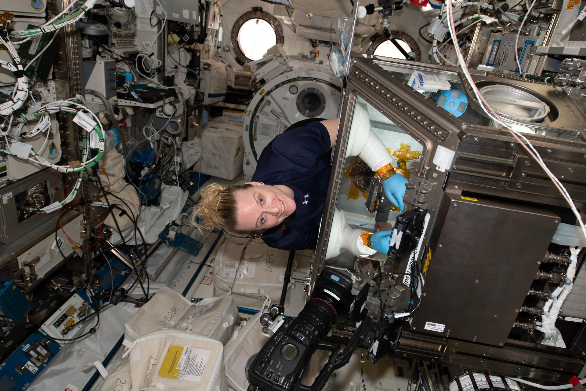 Rubins stands at an angle wearing a navy-blue shirt, smiling while conducting an experiment inside a glovebox. The metallic glovebox is about the size of a washing machine, with a glass cover. Rubins’ arms are inserted through the glass cover, wearing protective blue latex gloves and white protective sleeves. A black camera is pointed towards the glovebox, and the surrounding walls have many wires, hardware, and containers attached.