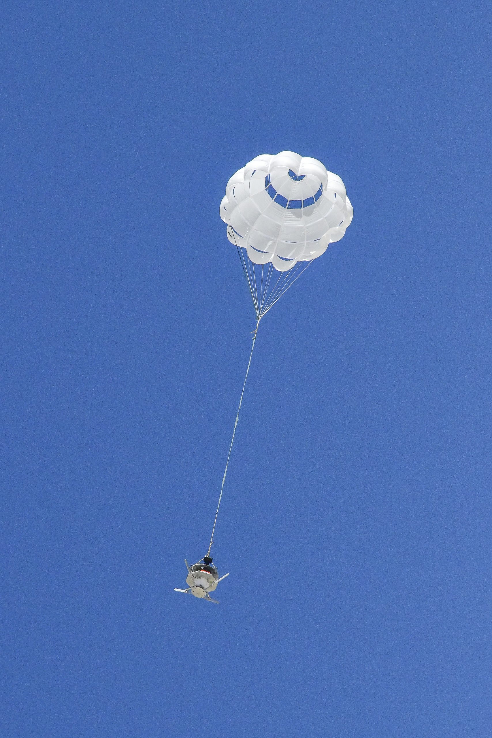 A parachute carrying a capsule is fully open against a canvas of deep blue sky.