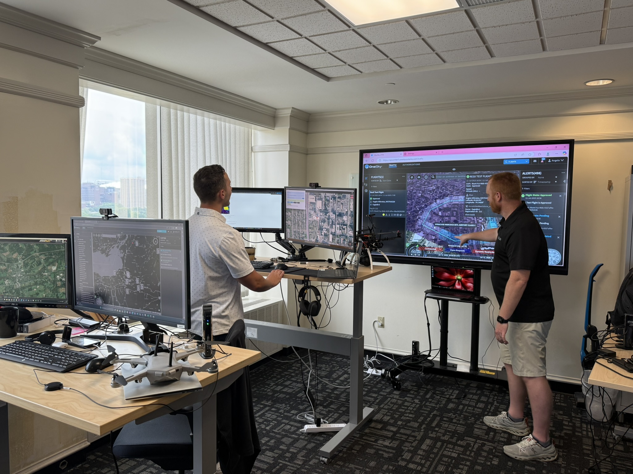 Two men stand in an office looking at four smaller computer screens and one larger computer screen. There is a tan desk in the foreground housing two computer monitors and a small grey drone. In the background stands a man in a white polo shirt facing two other computer monitors on a desk. Behind this set up is a large TV monitor with a man standing with his arm reached out in front. The man is wearing a black polo shorts and tan shorts as well as white tennis shoes. The screen shows a map and different pieces of software working in unison.