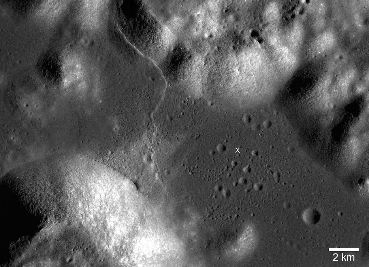 A grayscale image shows an uneven surface with various textures, including smooth areas, ridges, and depressions. The surface features several small, round indentations, as well as larger, irregularly shaped mounds and valleys. A thin, winding line crosses diagonally through the image, resembling a fissure or channel. A small white "x" marks the center of the image. A scale bar in the bottom right corner indicates 2 km.