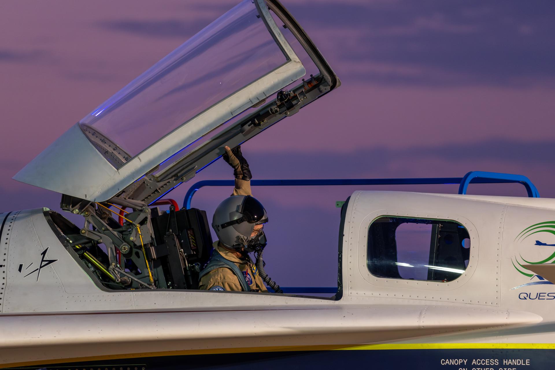 NASA’s X-59 lead test pilot Nils Larson sits in the cockpit of the X-59 wearing a helmet and tan flight suit under a green G-suit. The X-59’s canopy is partially raised, and Larson can be seen reaching up with his left hand to close it.  The Quesst mission logo is visible on the aircraft’s fuselage and the background sky has a soft purple and pink color from the early light.