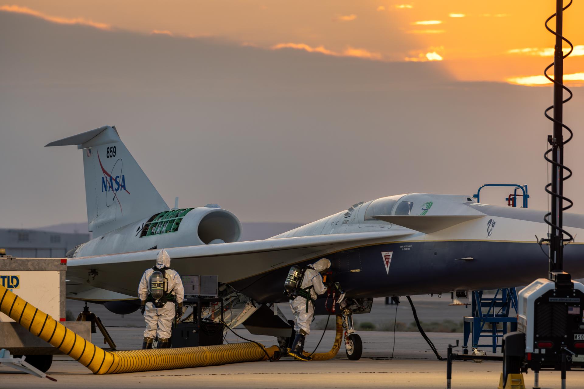 Two maintainers in white protective suits, air tanks, masks, rubber gloves, and boots check NASA’s X-59 hydrazine system during a hydrazine safety check at U.S. Air Force Plant 42 in Palmdale, California. The sky is slightly overcast, with some sunlight rays beginning to peek out from behind the clouds. The aircraft has a panel open, a ladder positioned on the right side of its fuselage, and air ducts extending from the aircraft into a service cart.