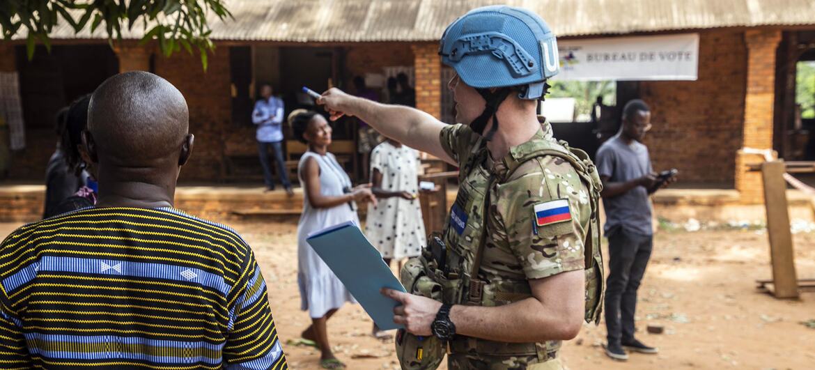 A UN peacekeeper in a blue helmet and military uniform points at a ballot box while overseeing an election at a 'Bureau de Vote' in Africa.