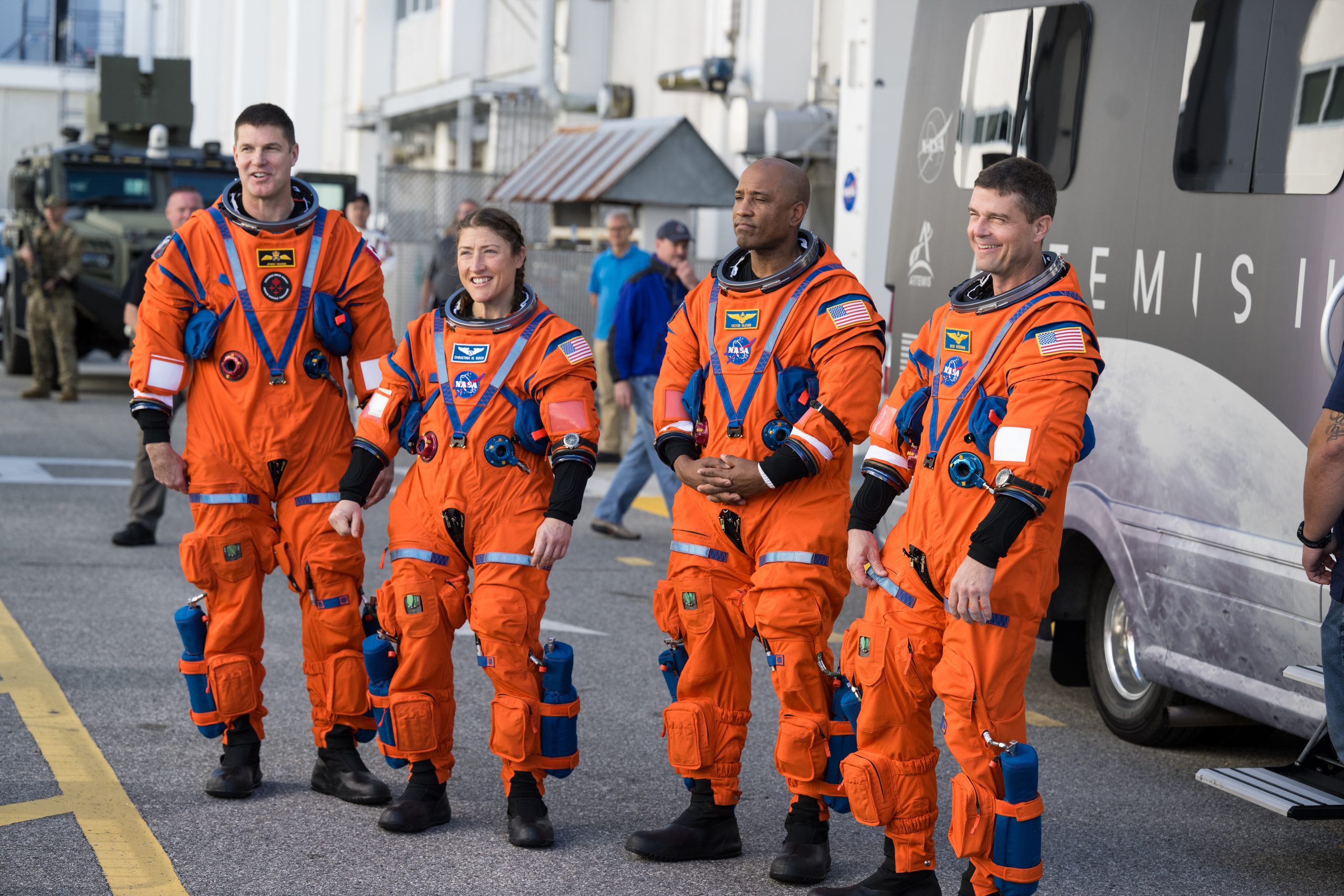 Four astronauts - three men and one woman - wearing orange spacesuits stand on the street. Behind them, people are standing or walking by. There is a partially visible van behind the crew; the van has a picture of the Moon and the words "Artemis II" on it.