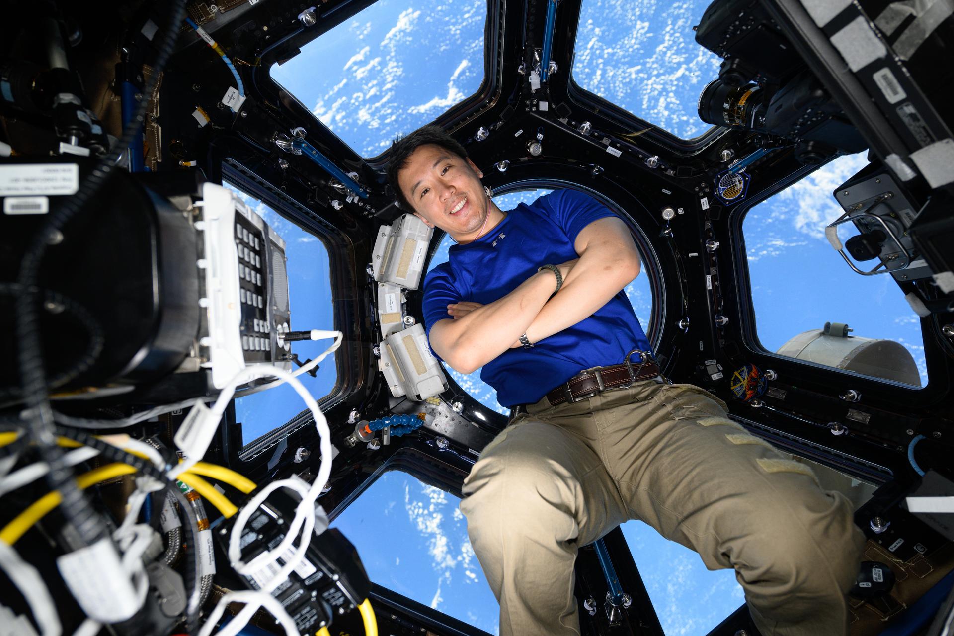 NASA astronaut Jonny Kim floats in the center of the image with his arms crossed, smiling. He wears a blue shirt and khaki pants. Behind him, the seven windows of the space station’s cupola reveal Earth’s blue oceans below. In the foreground, on the left, a rectangular device with a keypad and multiple wires is visible.