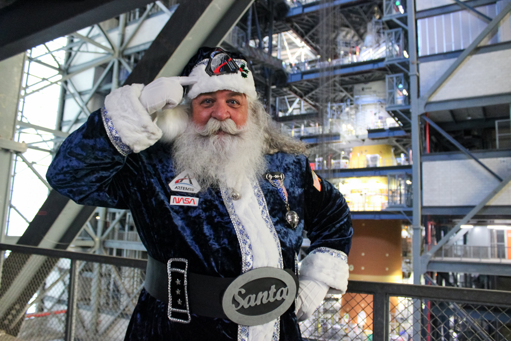 A man with a big white beard wearing a blue Santa costume with Artemis and NASA patches points to his hat. The Artemis II SLS and Orion spacecraft are In the background.