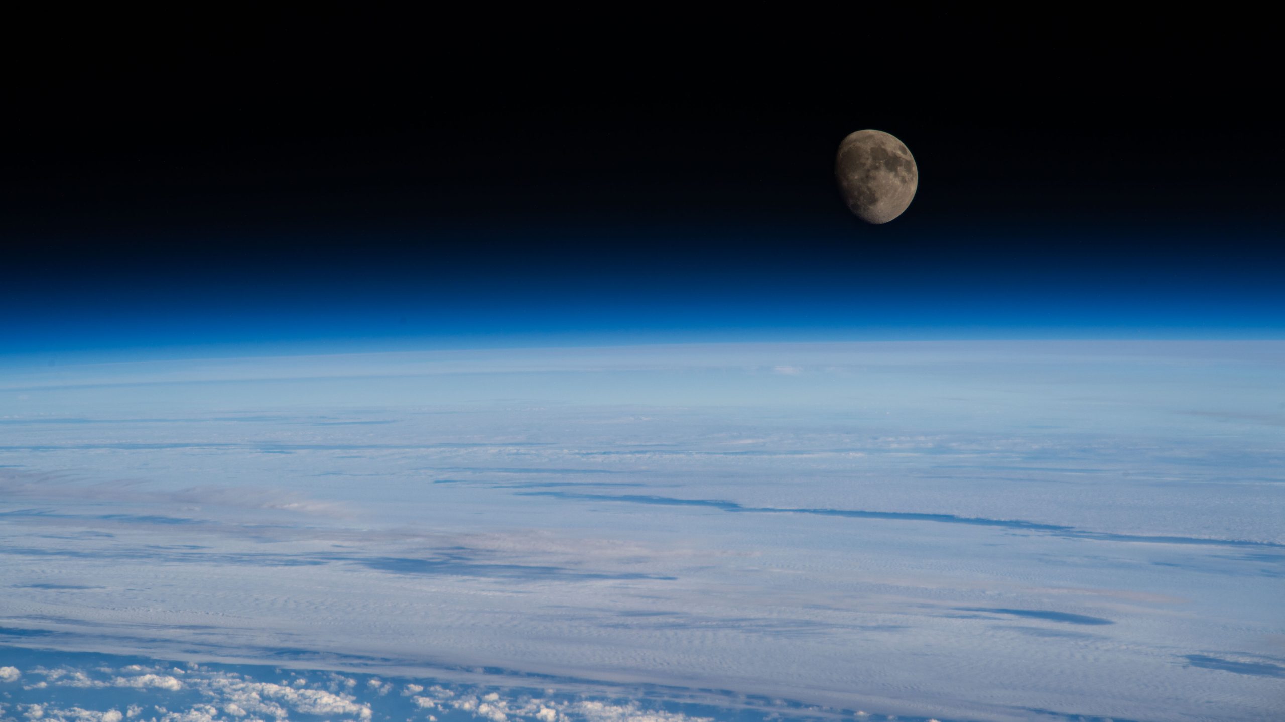 The Moon is nearly full and a deep gray-brown color in this image. Below it is Earth’s blue atmosphere and and white clouds. This photo was taken from the International Space Station.