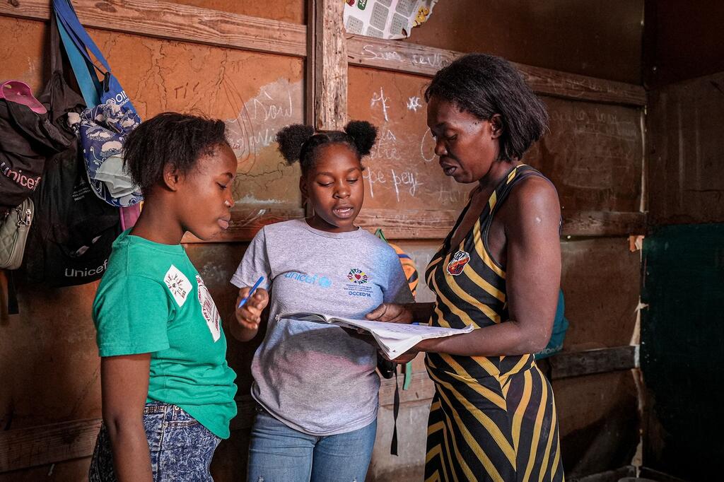 A young girl named Dieussika, wearing a UNICEF shirt, is being helped by her mother and a teacher while studying at a makeshift school in Port-au-Prince, Haiti. She has returned to education after displacement and is now graduating, highlighting the impact of UNICEF's educational support.
