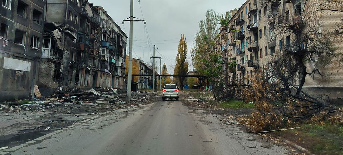 A white car drives down a damaged street in Ukraine, flanked by heavily bombed apartment buildings with broken windows and charred facades.