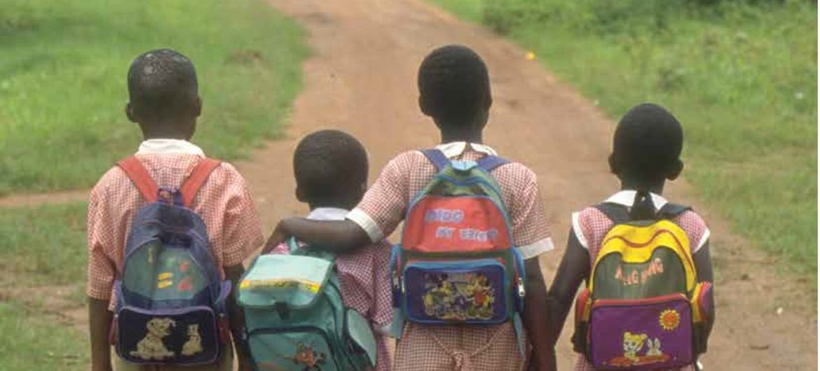 Four children in school uniforms walk down a rural dirt path with backpacks, two of them walking arm in arm.