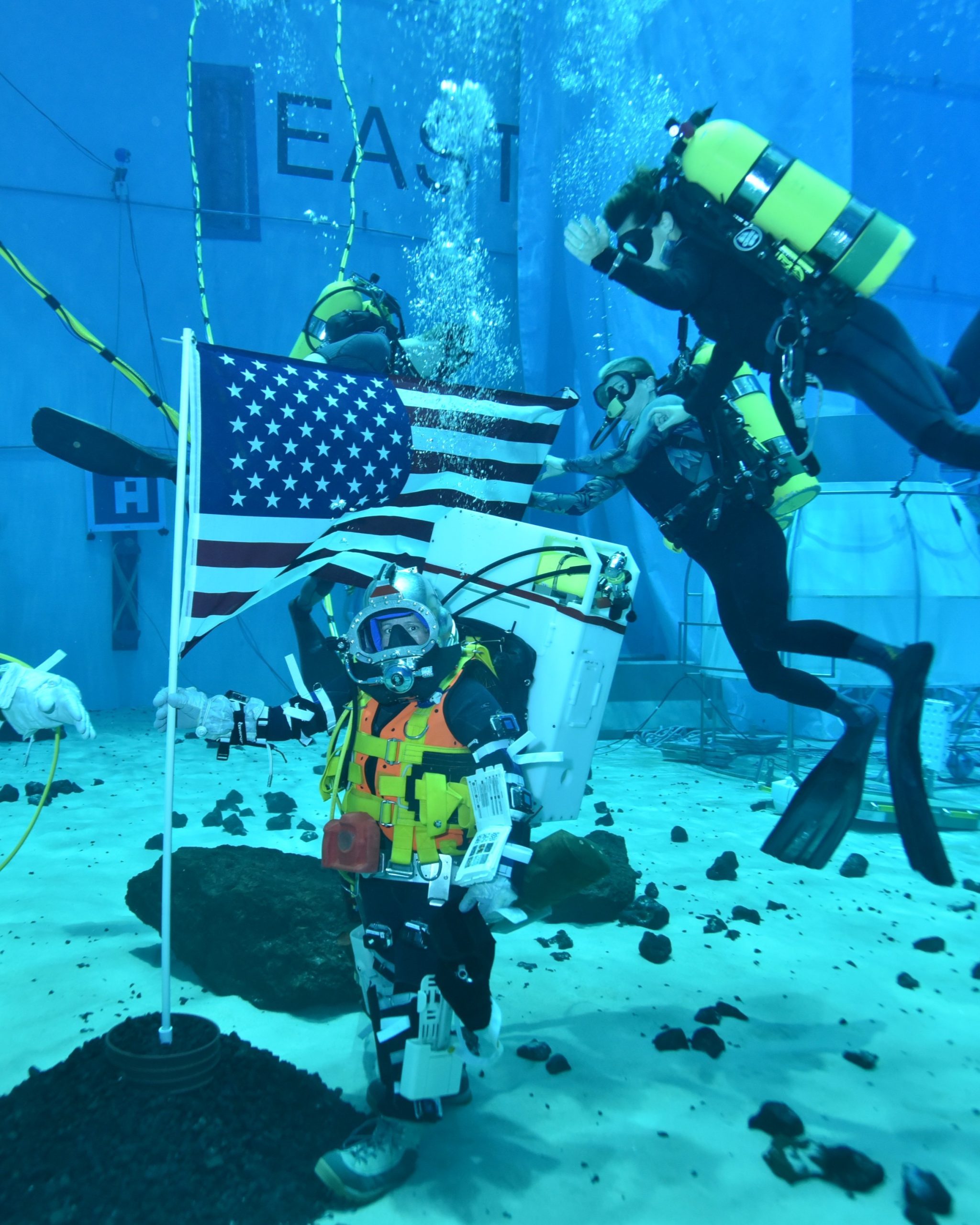 Two people practice underwater operations in a Moon like environment. The person on the left is holding an U.S. flag.