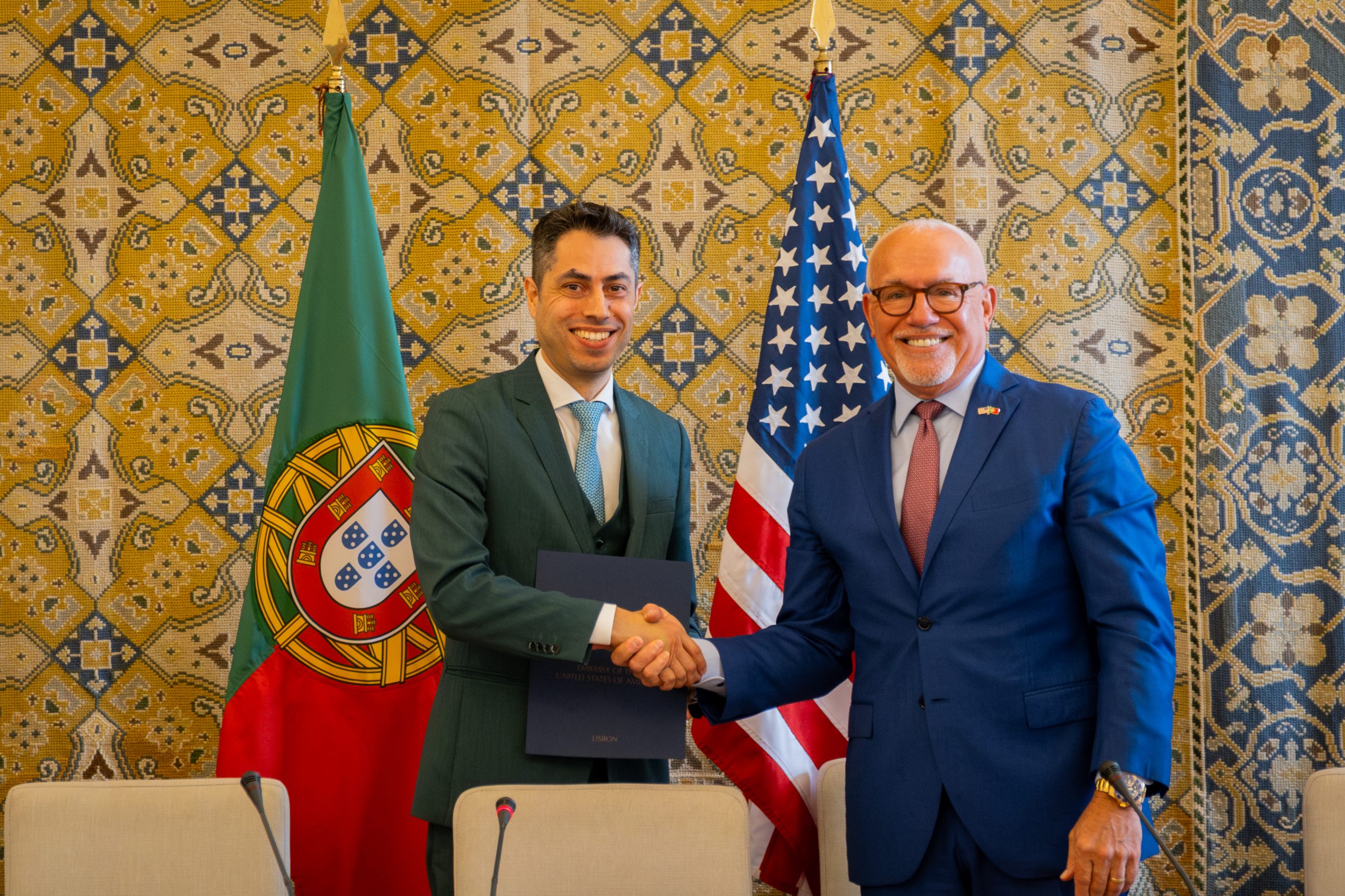 Hugo Costa, executive director for Portuguese Space Agency, and U.S. Ambassador to Portugal John J. Arrigo, shake hands and pose for a photo with country flags in background.