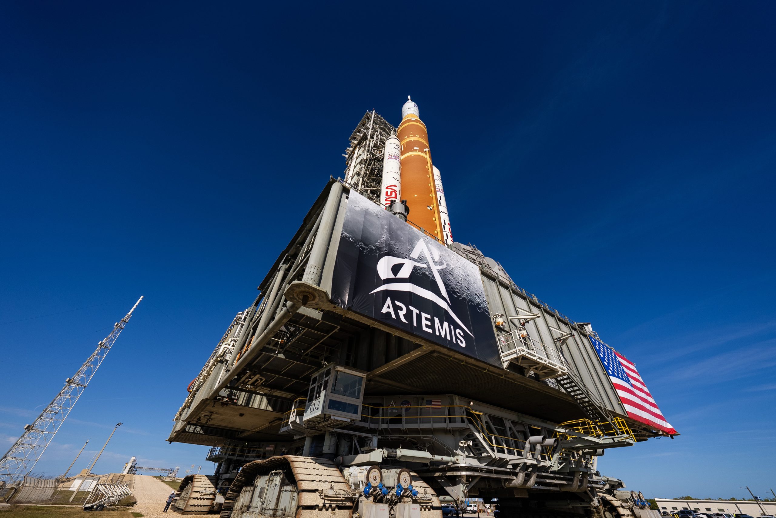 NASA's Artemis II SLS rocket - with its distinctive butterscotch orange-brown color- and Orion spacecraft are on top of a massive platform. The platform has a banner with the Artemis logo on it on the left and an American flag on the right. The rocket and platform are moved by an immense machine called the crawler-transporter, which has treads like a tank. The view is from the ground up, so the deep blue sky is the background for most of this picture.