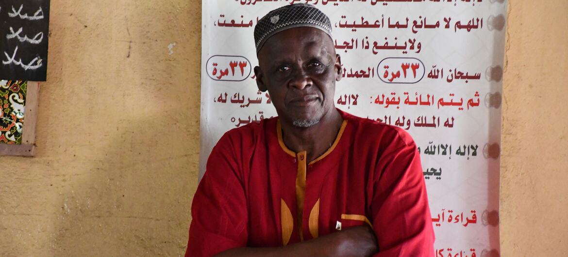 A man in a red shirt and headwrap stands with arms crossed in front of a wall with Arabic text, symbolizing community resistance against female genital mutilation in Guinea.