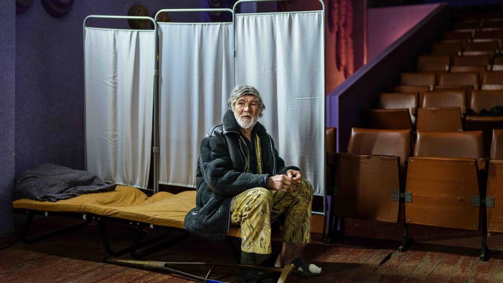 Elderly Ukrainian refugee sits in a theater-turned-shelter amid ongoing war. An elderly Ukrainian man sits on a cot in a theater-turned-shelter, surrounded by rows of empty seats and privacy screens, highlighting the humanitarian crisis caused by the ongoing war.