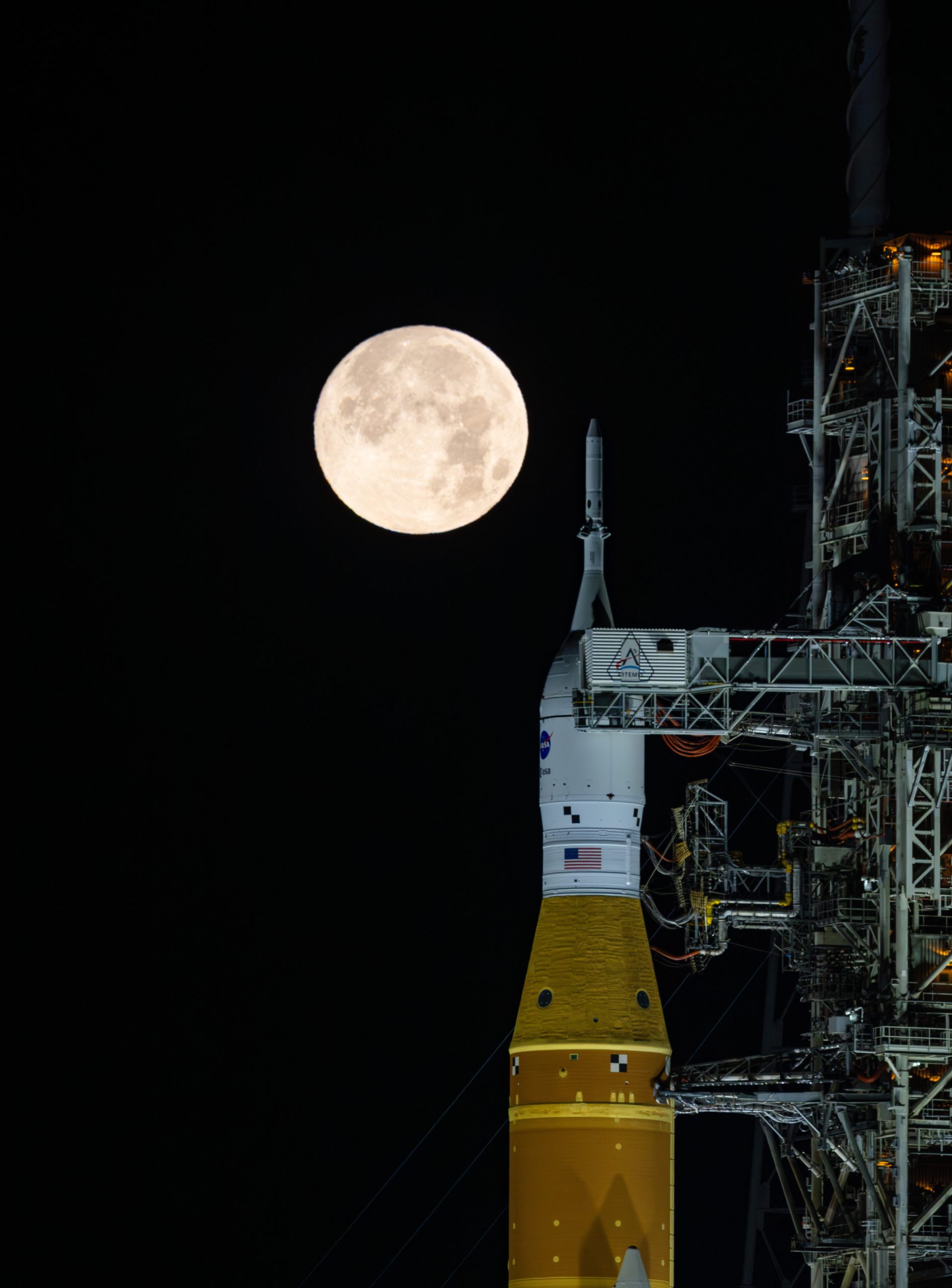 A golden full moon is seen shining over NASA’s SLS (Space Launch System) and Orion spacecraft, atop the mobile launcher in the early hours of February 1, 2026. The SLS core stage is a distinctive deep orange, while Orion is white and sits on top of the core stage.