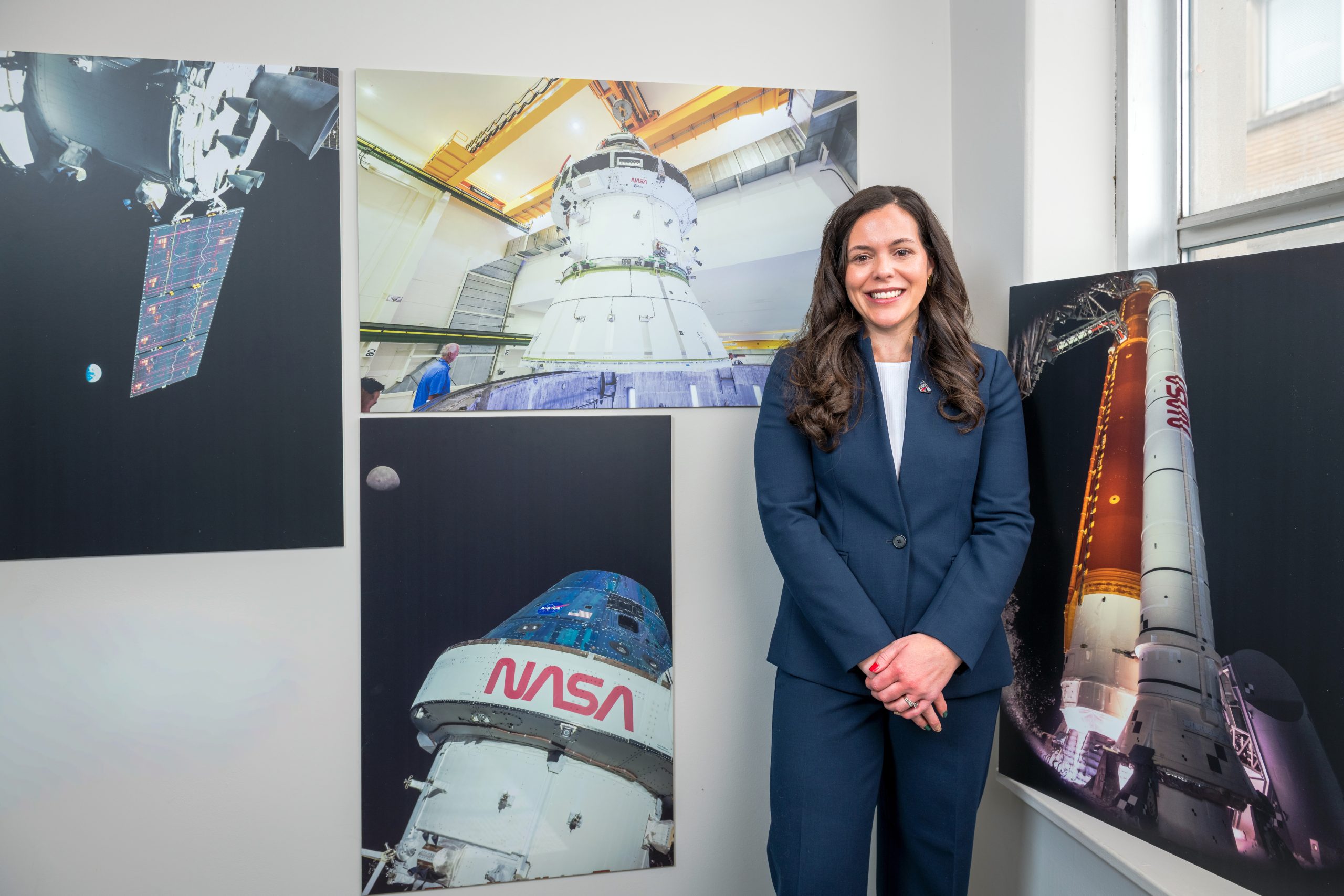 A professionally dressed woman stands in a gallery style room surrounded by large NASA photographs showing the Orion spacecraft in space, engineers working on a module, a close-up of the Orion spacecraft with the Moon behind it, and the SLS (Space Launch System) rocket illuminated on the launch pad.