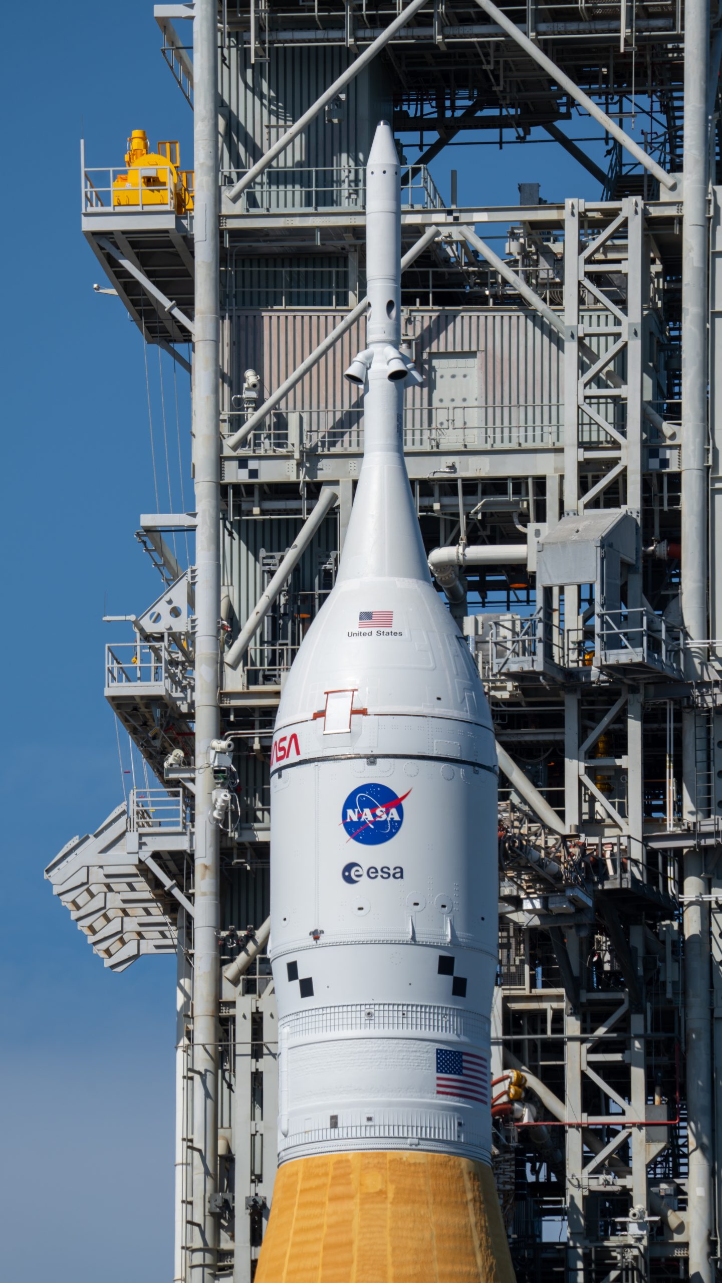 The Orion spacecraft is white and cylinder-shaped with a pointed nose at the top. It rests above the SLS rocket, part of which (the orange part) can just barely be seen at the bottom of the photo. Behind Orion and the SLS is a gray gantry with scaffolding and balconies.