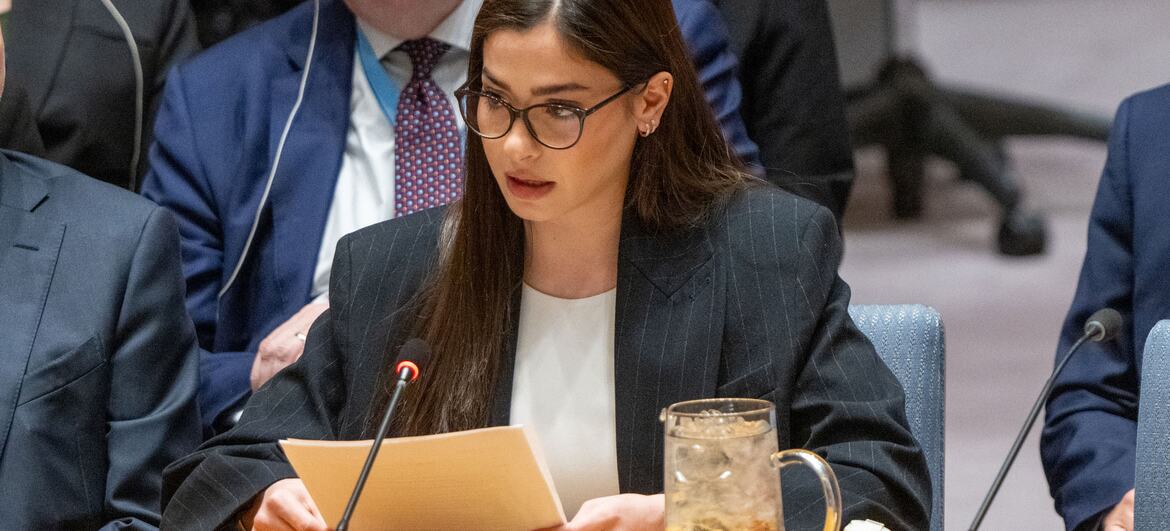Yusra Mardini speaking at a formal event, seated at a table with a microphone.