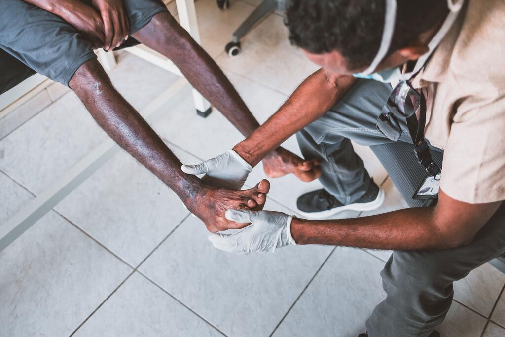Jamal is treated for his injuries at a shelter for migrants in Djibouti. A medical worker wearing gloves examines the injured foot of a seated African migrant in a clinic setting.