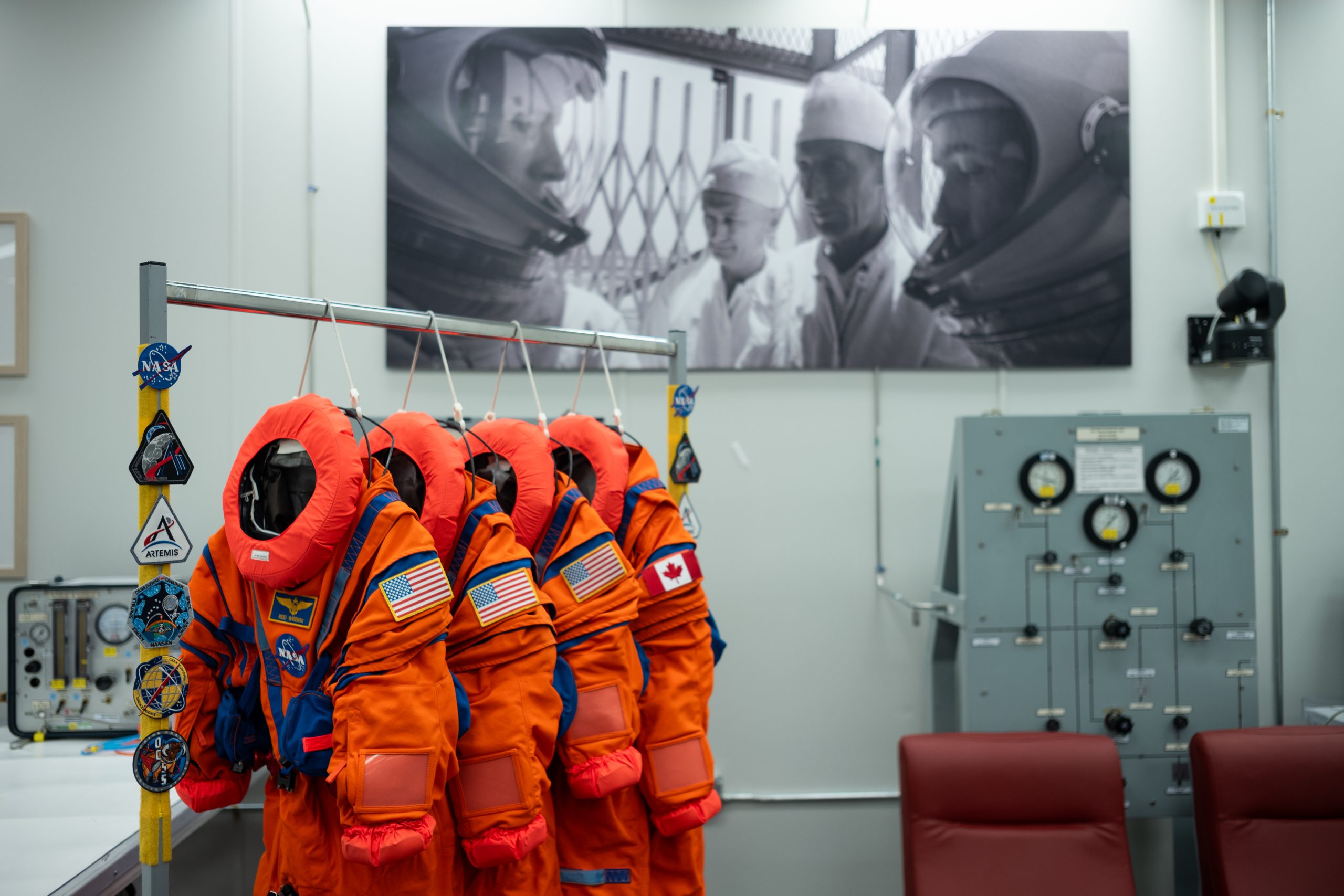 Four neon orange spacesuits hang on a rack decorated with NASA patches. Three of the suits have American flag patches on their left shoulder, while the last has a Canadian flag patch on its left shoulder. The room the suits are hanging in has a large black and white photo on the wall, in which astronauts are in their spacesuits and helmets while two people in white lab coats look on.