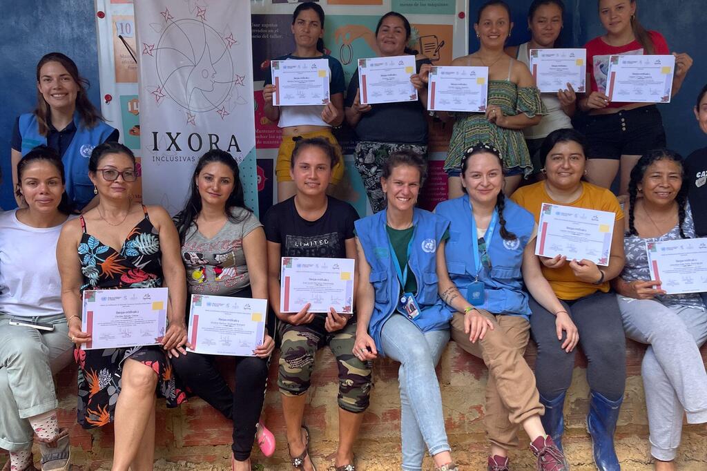 Women from the Ixora brand at the trade show following the conclusion of a garment-making workshop organized by UNVMC A group of women, including UN Verification Mission in Colombia staff and fashion designers, pose together holding certificates. They are smiling and standing in front of an IXORA Inclusive banner.