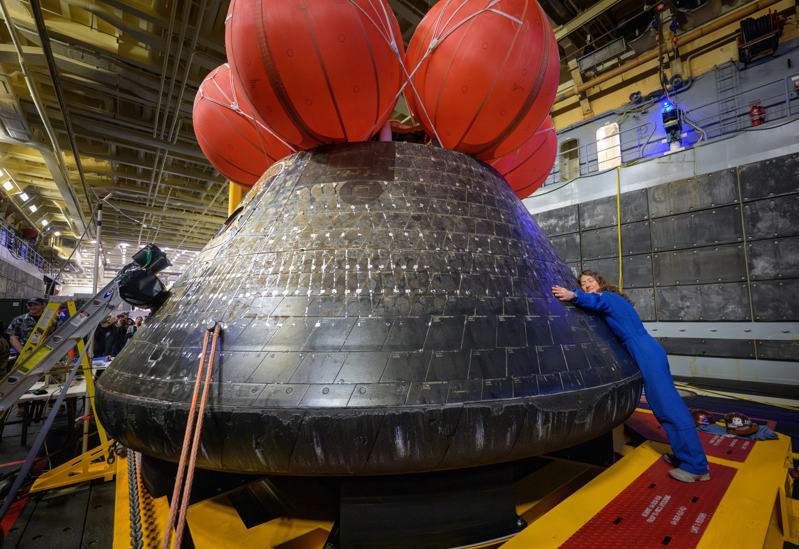 NASA astronaut Christina Koch hugs the Orion spacecraft with her face toward the camera. She is wearing a bright blue jumpsuit. Orion is a rounded cone shaped spacecraft. Its exterior is a sooty black. There are several large orange balloons attached to its top. Orion looks about twice Koch's height.