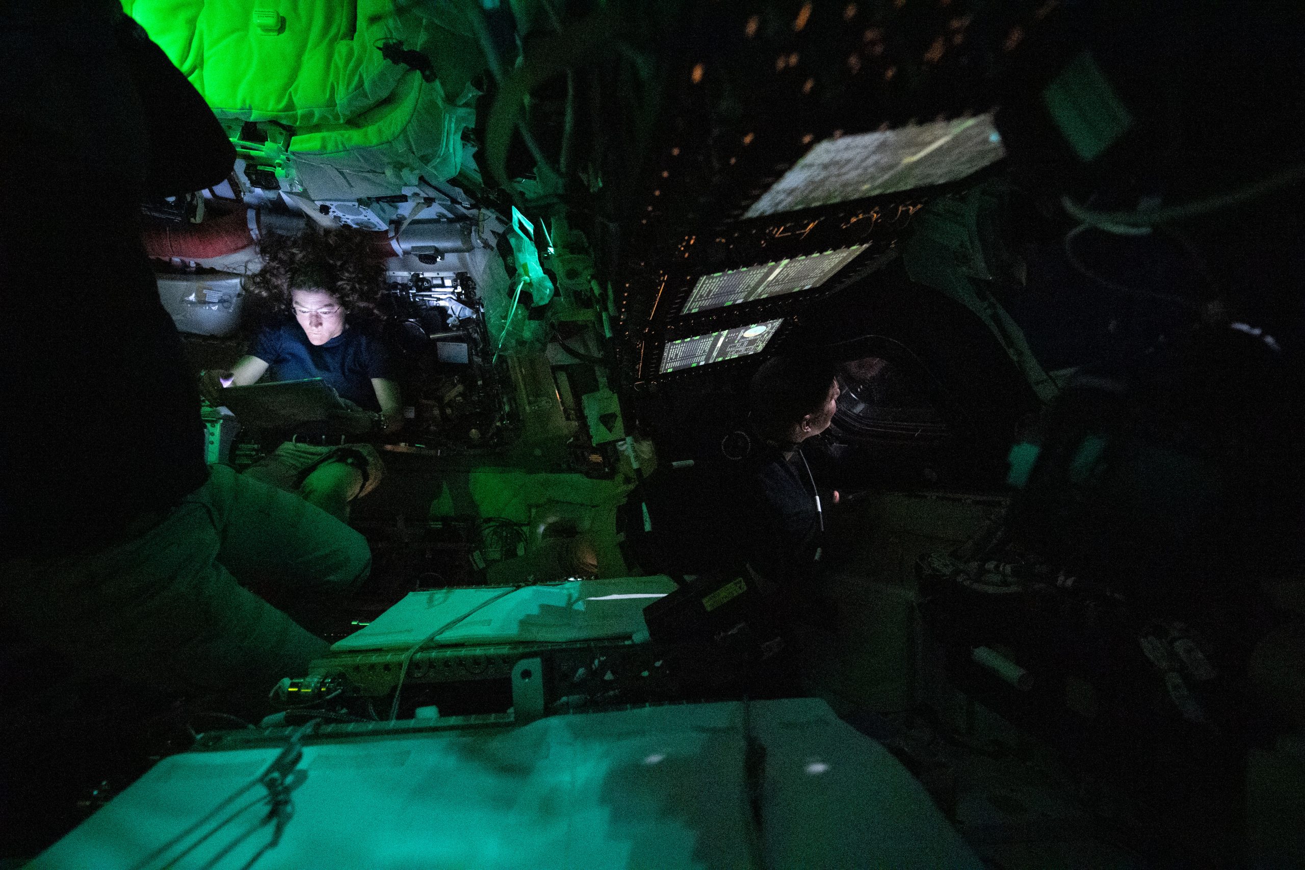 Astronaut Christina Koch is nestled into a corner of the Orion crew capsule, reading. The rest of the capsule is dark, but her face is lit by the glow from her tablet. Two of her other crew members can dimly be seen looking out the window and floating in the foreground.