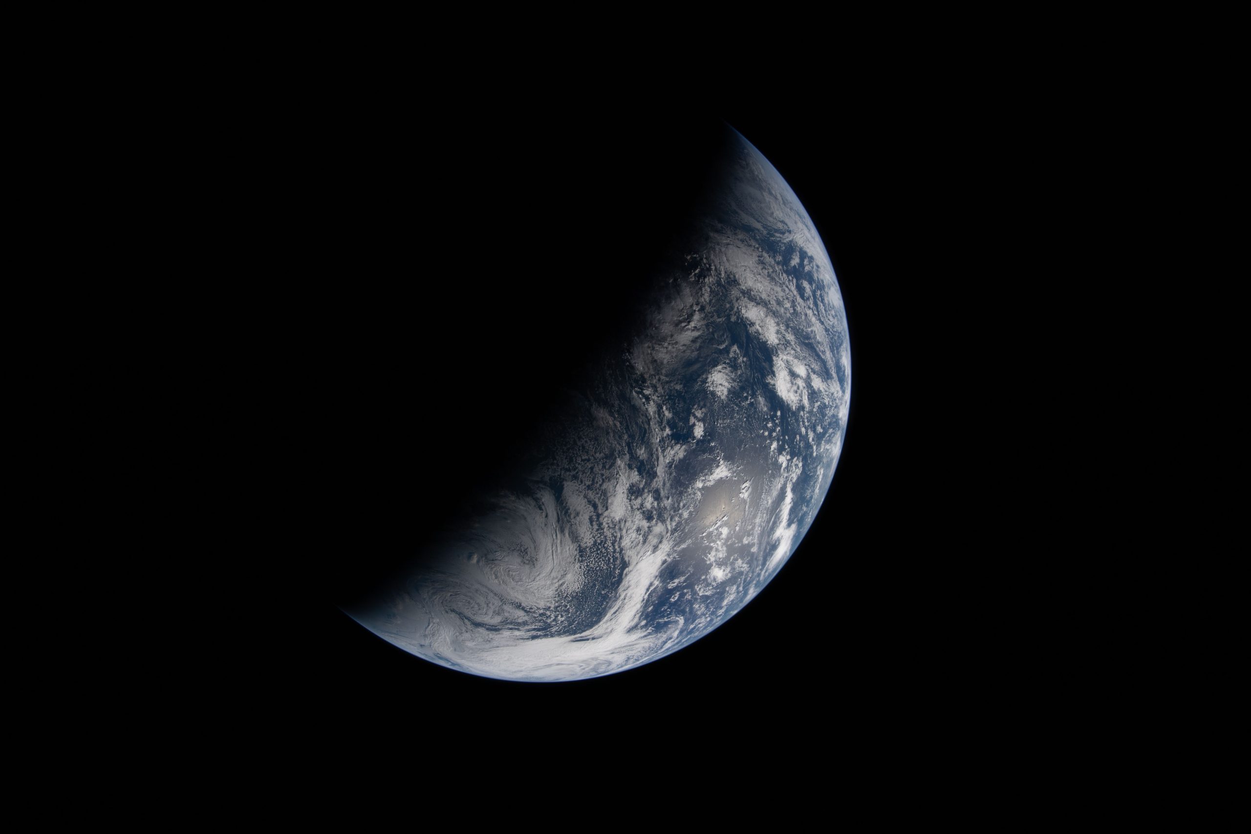 Image of Earth split by the terminator line. The terminator divides Earth’s surface diagonally, shrouding the top left half in darkness. Earth’s illuminated surface is mostly blue and covered in swirling white clouds.