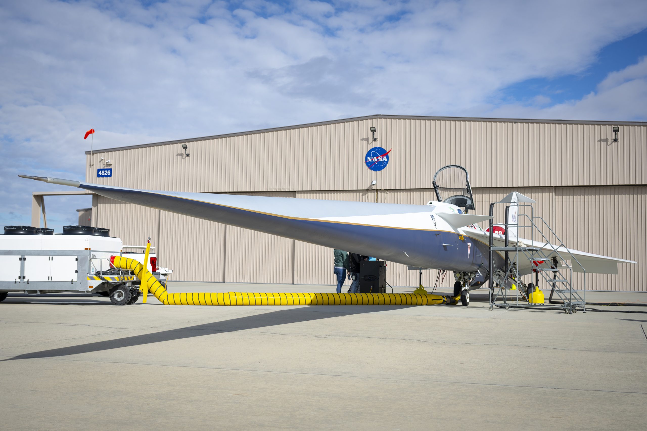 A white and blue jet airplane is parked in front of a building with large sliding doors and a NASA logo centered on the forward wall. The building is the new X-59 hangar.