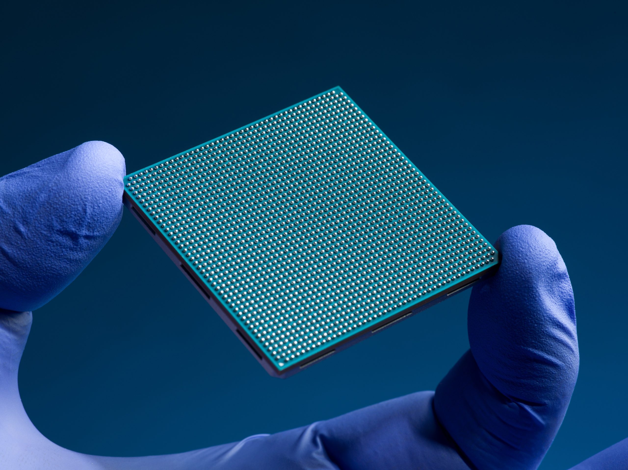 The finger and thumb of a hand clad in a blue nitrile glove hold a teal-colored square semiconductor chip against a dark blue background. The chip has an intricate grid of hundreds of tiny silver solder bumps on its underside.